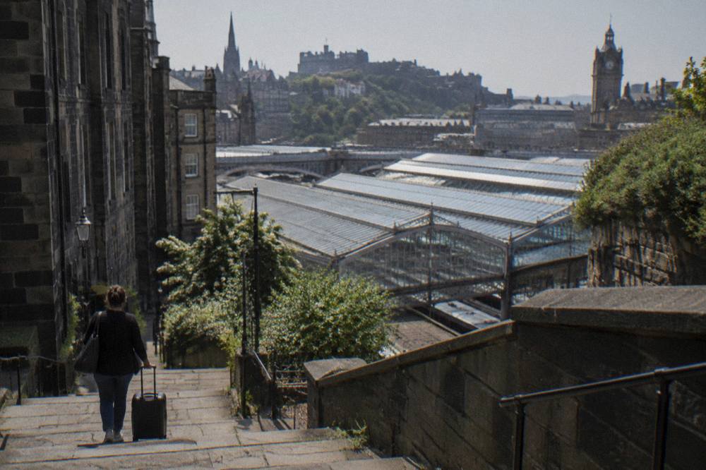 A traveller with a rolling suitcase descends stone stairs toward a glass-roofed station and castle skyline