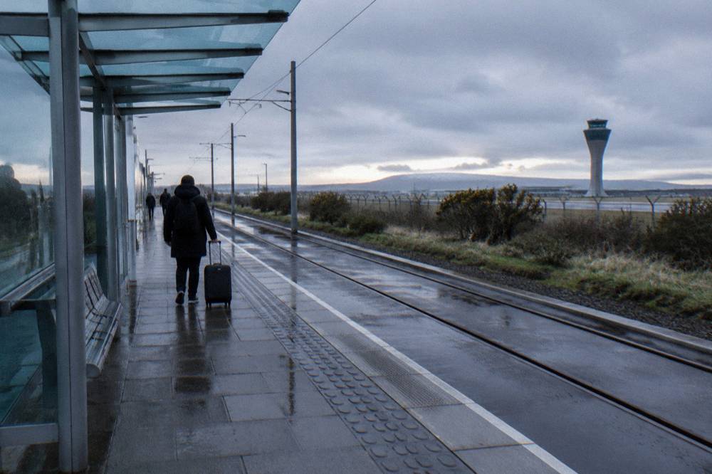 Person pulling a suitcase at a wet tram stop near an airport control tower