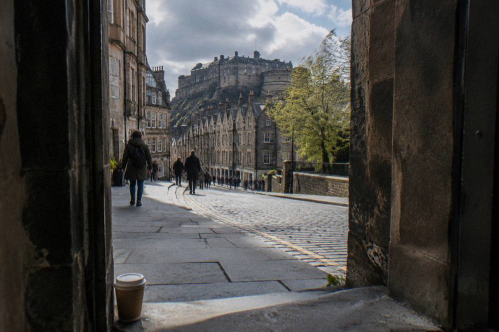 A cobblestone street viewed through an arch, with pedestrians and a castle atop a hill