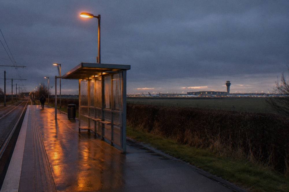 A bus shelter beside a road at dusk with an airport terminal and control tower in the distance