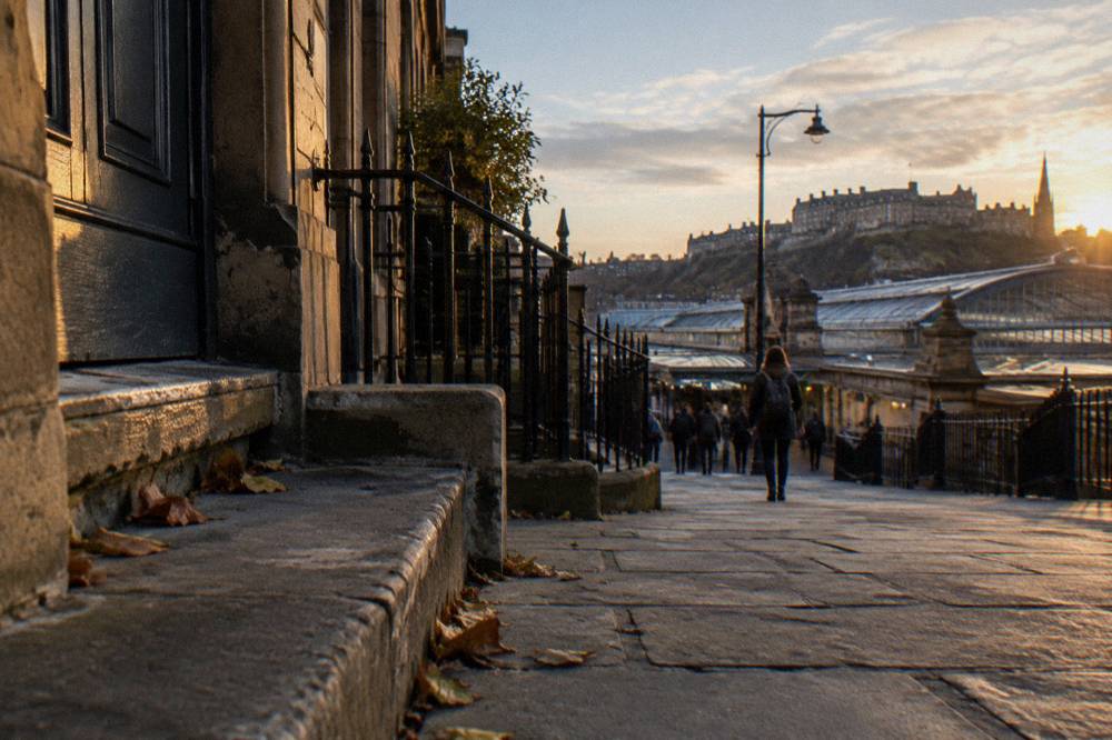 Stone steps and iron railings beside a doorway on a cobbled street, castle silhouette at sunset