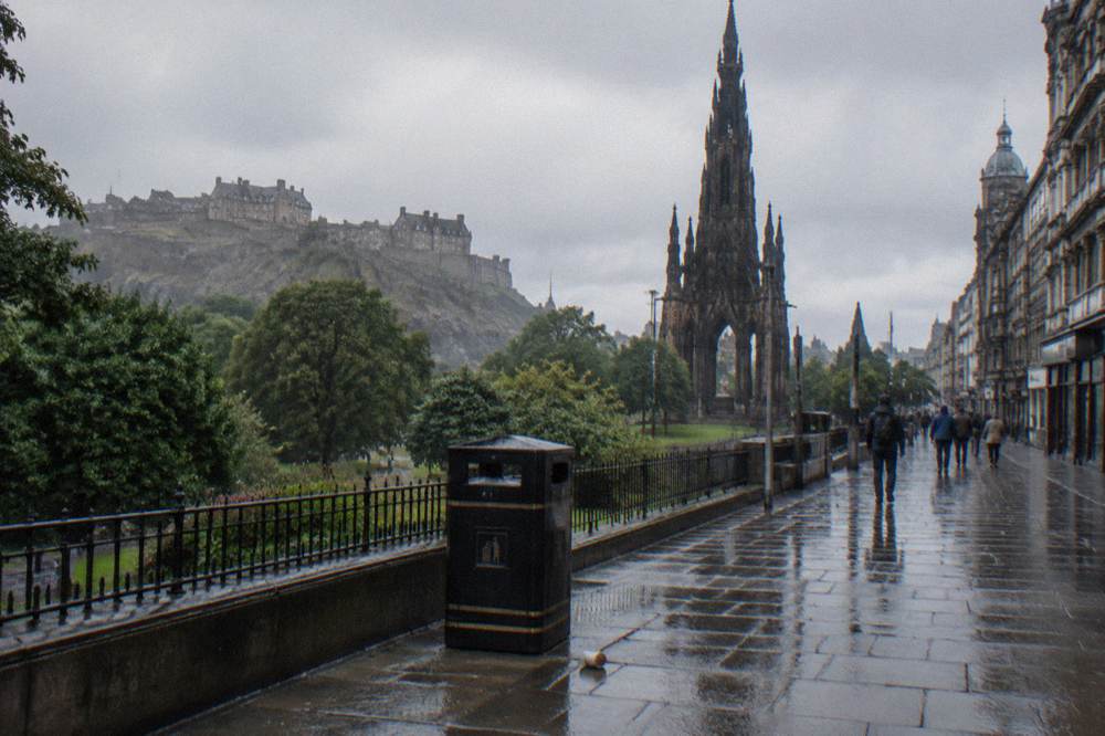 Rainy stone promenade with ornate gothic tower monument, castle on hill, pedestrians, litter bin, overcast sky