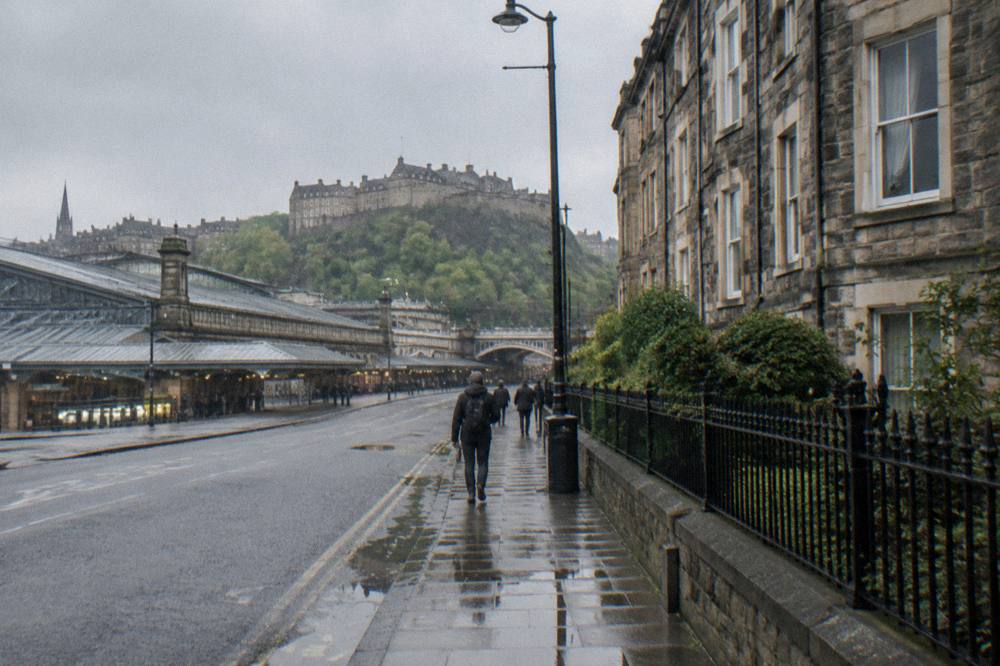 A rainy city street with pedestrians, stone buildings and a castle on a hill