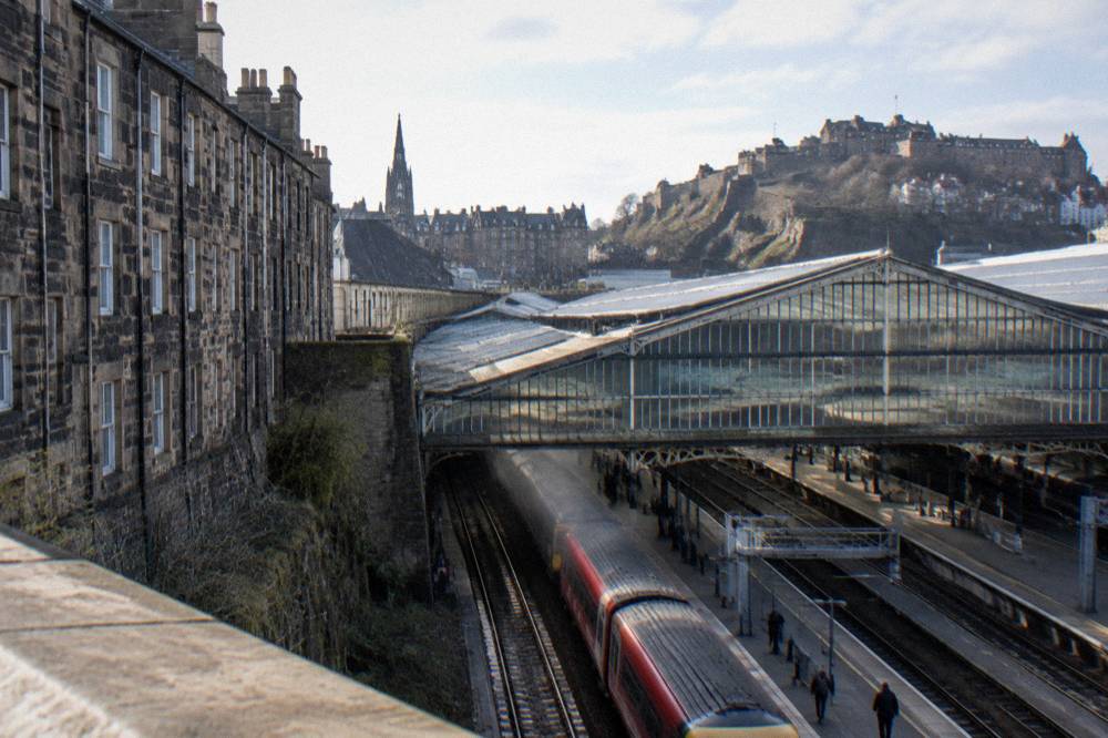 Railway station platforms with a glass roof, a train on tracks, and a castle on a hill