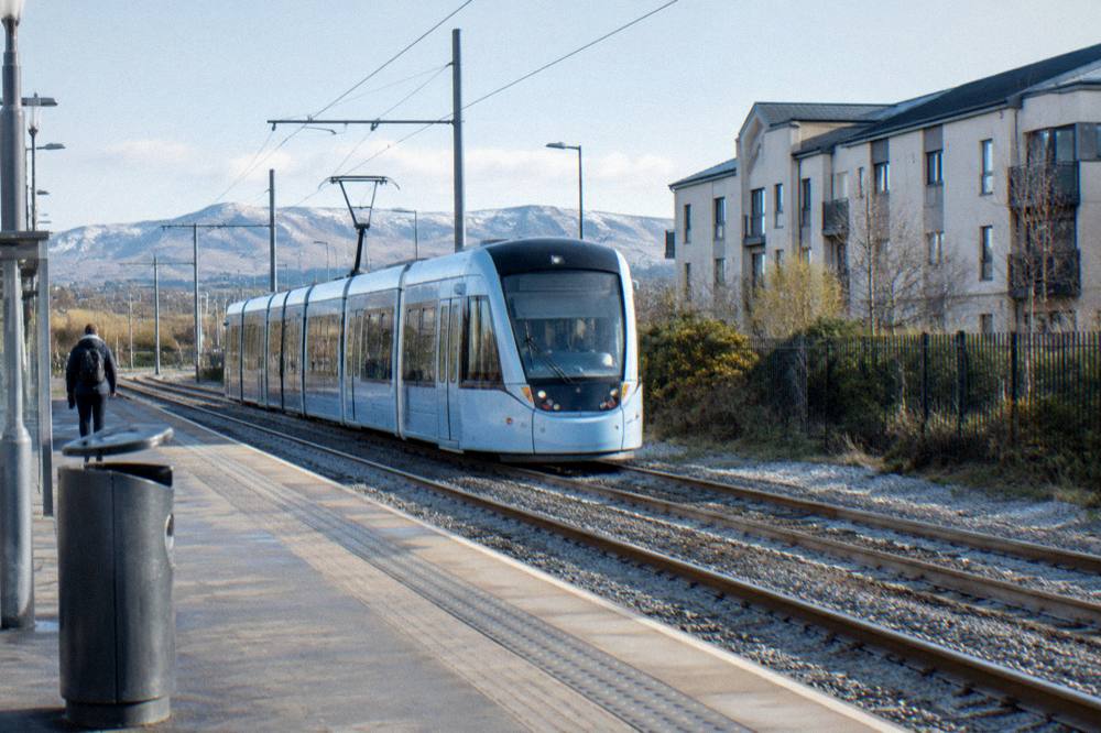 Tram passing modern apartment buildings on an Edinburgh street with a hill behind
