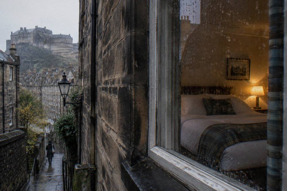 A cosy bedroom seen through a rainy window overlooking a wet alley with a castle on a hill