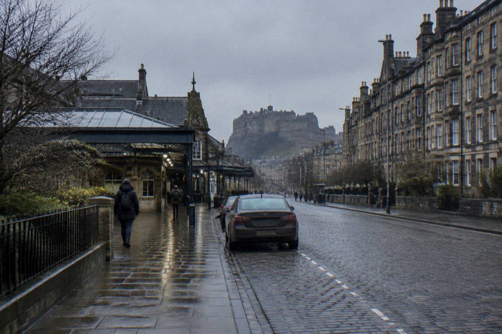A wet cobbled Edinburgh street with a parked car and castle in the distance