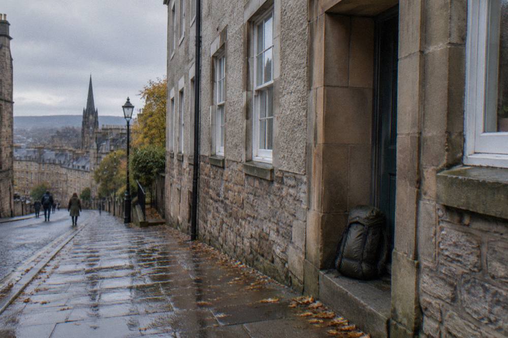 A wet stone street with old buildings, a church spire, pedestrians, and a backpack on a doorstep