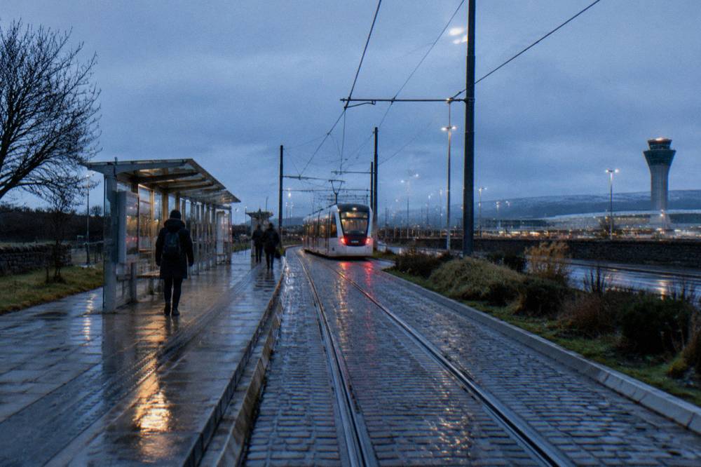 A tram approaches a sheltered stop on wet tracks, with an airport control tower behind