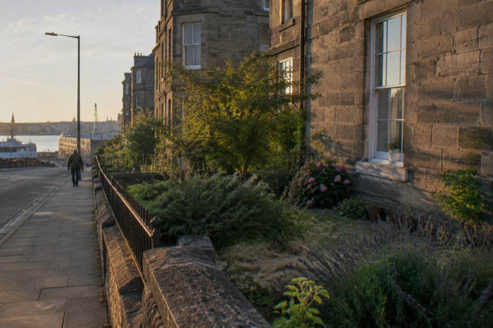 Stone terraced houses line a quiet pavement at sunset, overlooking water with a distant ship
