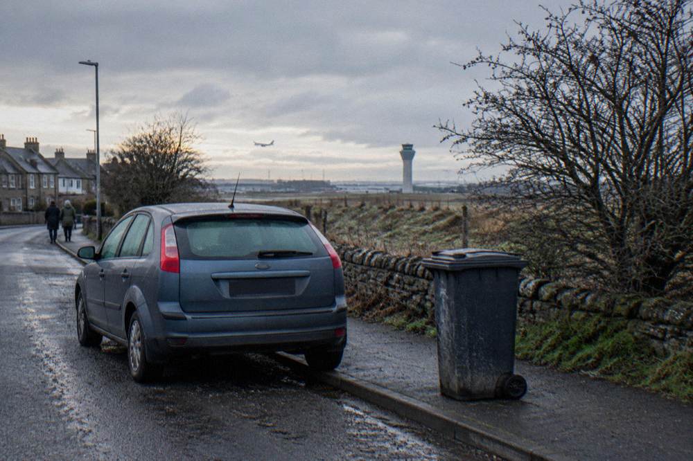 Grey car parked by a wheelie bin on a residential street, airport control tower and plane in distance