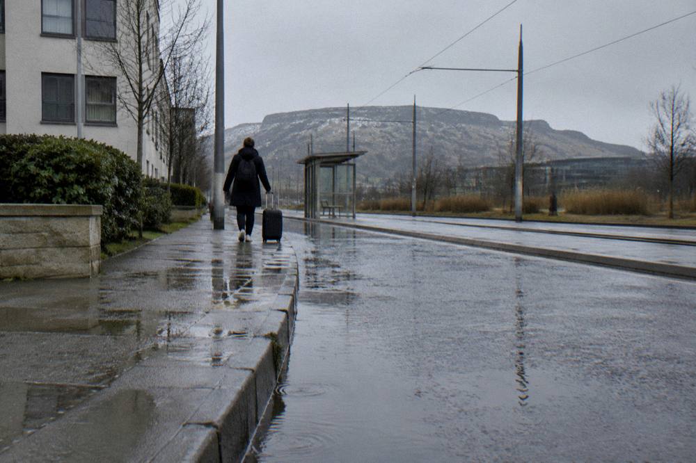 A person pulling a suitcase along a wet pavement beside tram tracks with a hill behind