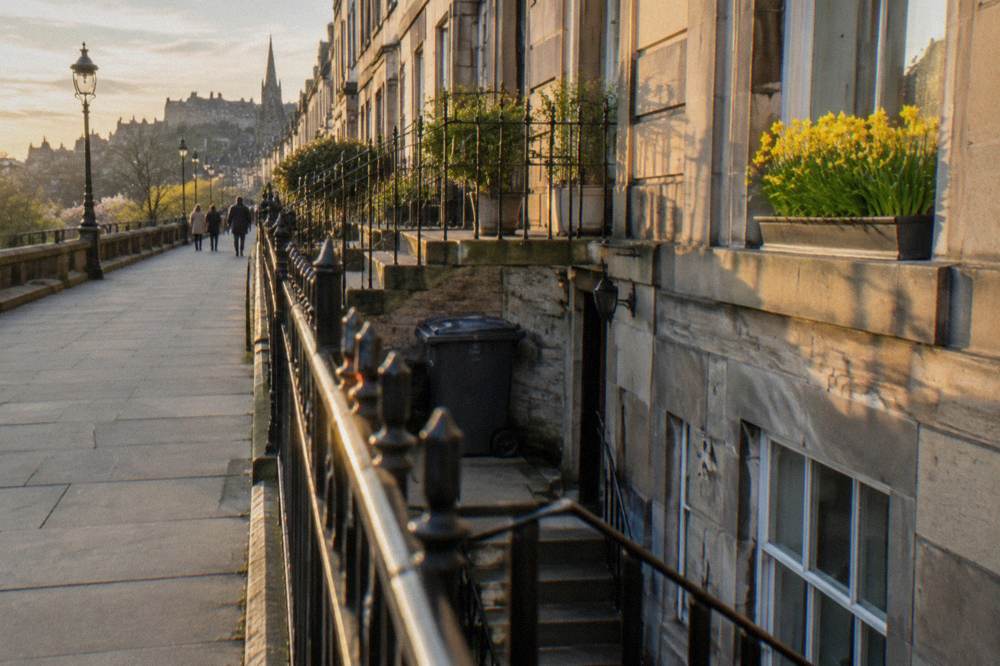 Sunlit sidewalk beside Georgian townhouses with iron railings and a distant castle skyline