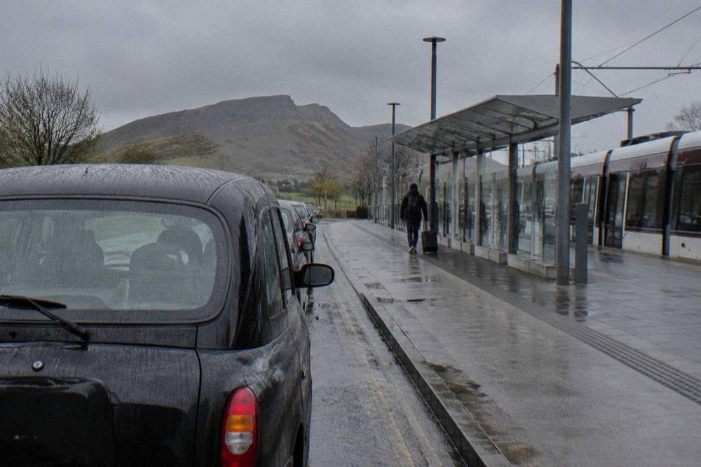 A black cab parked by a wet tram stop, with a tram, pedestrian, and hill behind