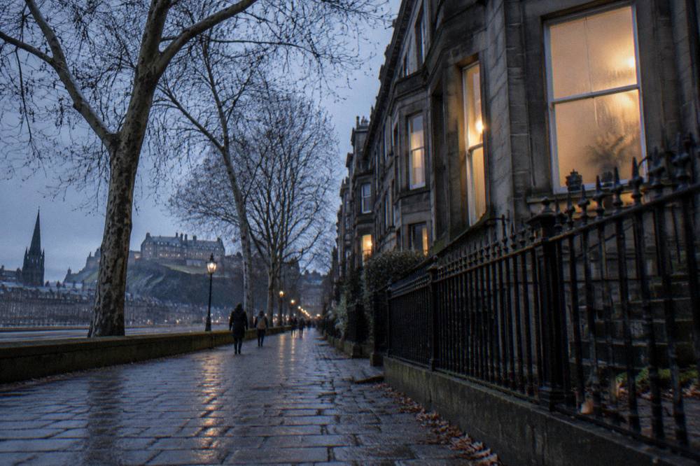 Exterior dusk street scene with wet cobbles, lit townhouse windows, bare trees and distant castle