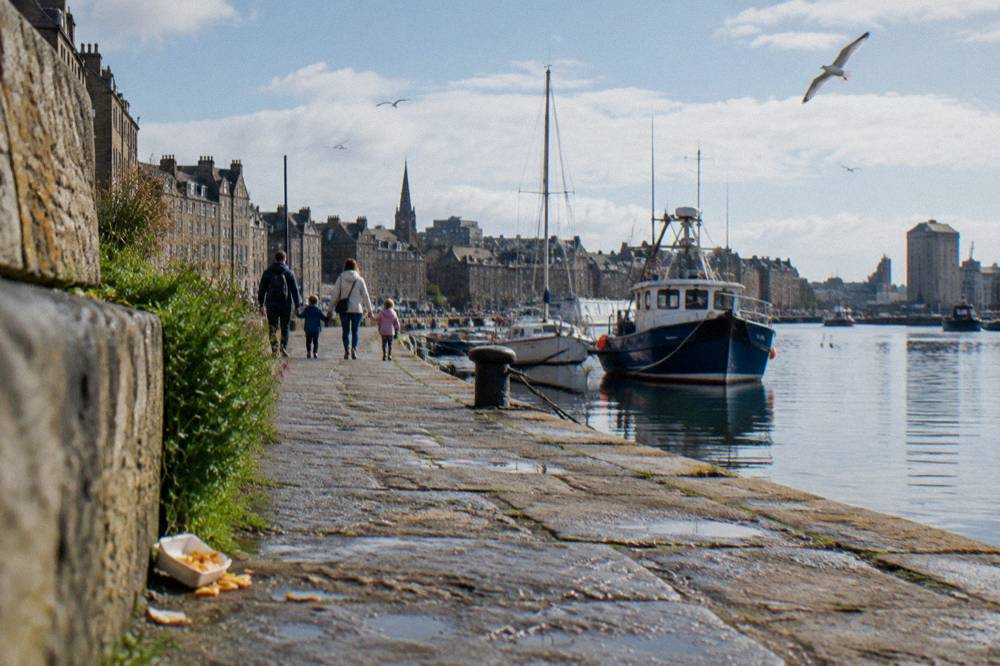A family walks along a cobblestone harbor quay beside moored boats and seabirds