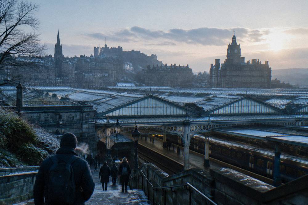People descending snowy steps toward a glass-roofed railway station at dusk, castle skyline behind