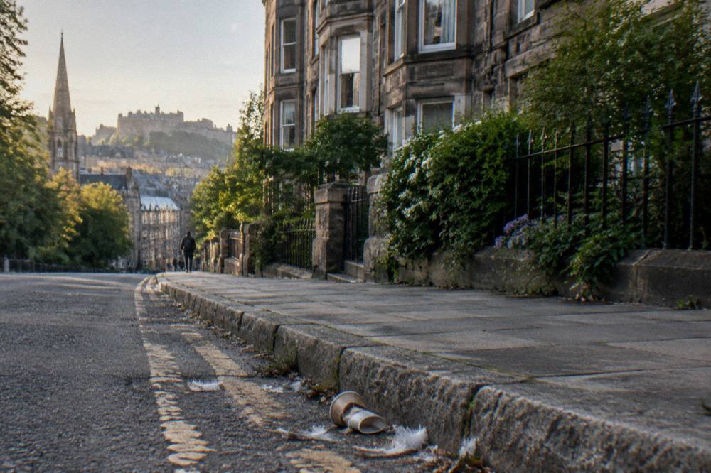 A sloping residential street with stone townhouses, a church spire, kerbside litter, and a castle skyline