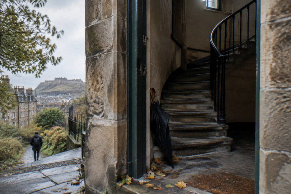Spiral stone staircase at a building entrance with a hanging umbrella and a wet street outside