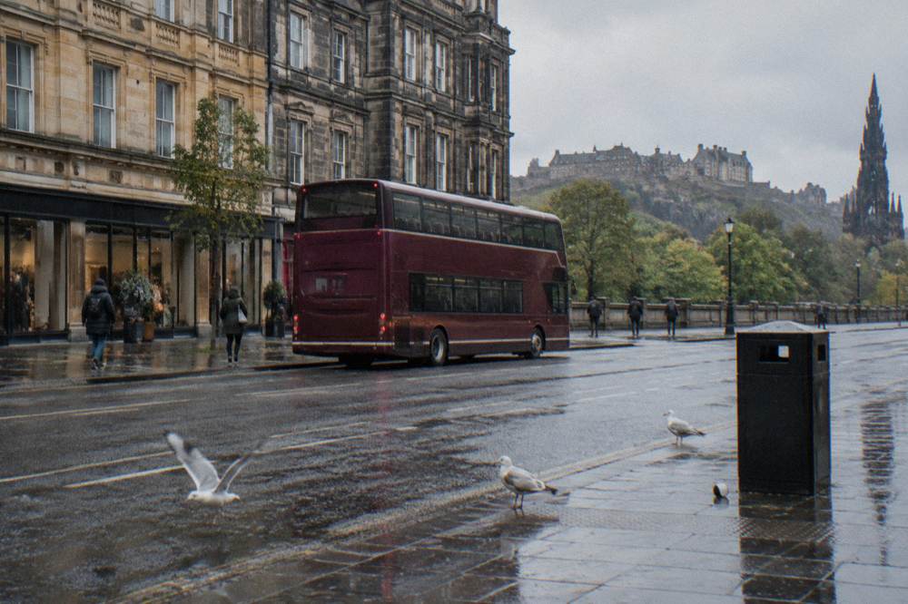 A red double-decker bus on a wet Edinburgh street with shops and castle behind